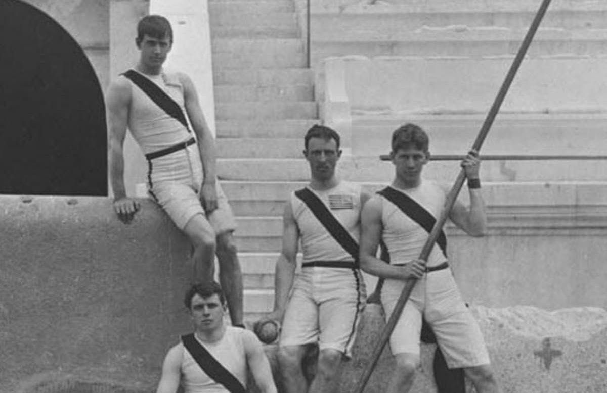 Four men in track and field uniforms pose for a photo. The photo is black and white.