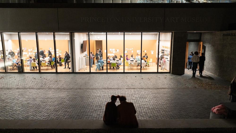 Two rooms are lit up in the darkness with people sitting around tables in the rooms. In the foreground, a dark terrace with two people sitting on the edge.
