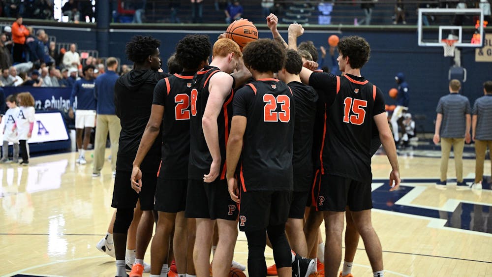 A group of Princeton players in black jerseys with orange letters and numbers huddle in a circle with their fists raised in the center.