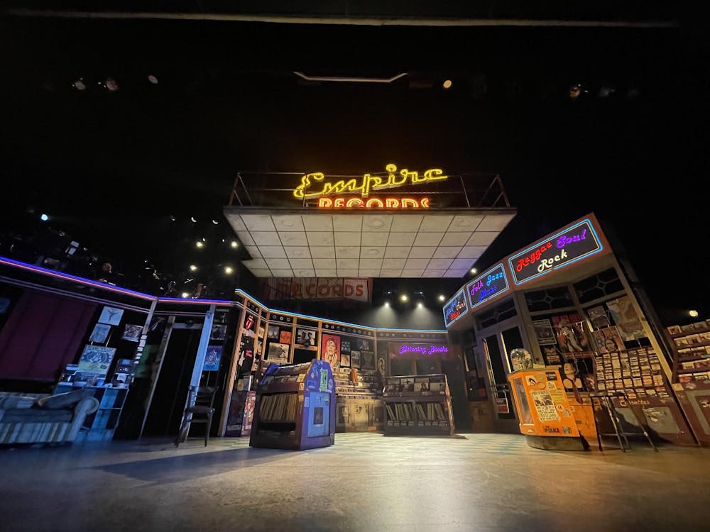 A stage production set up for a musical, with neon lights, vintage posters on walls, and shelves of vinyls.
