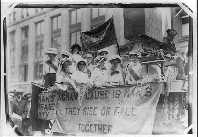 Suffragettes on a float in Yonkers, New York in 1913 with a banner reading "WOMANS CAUSE IS MANS / THEY RISE OR FALL"
