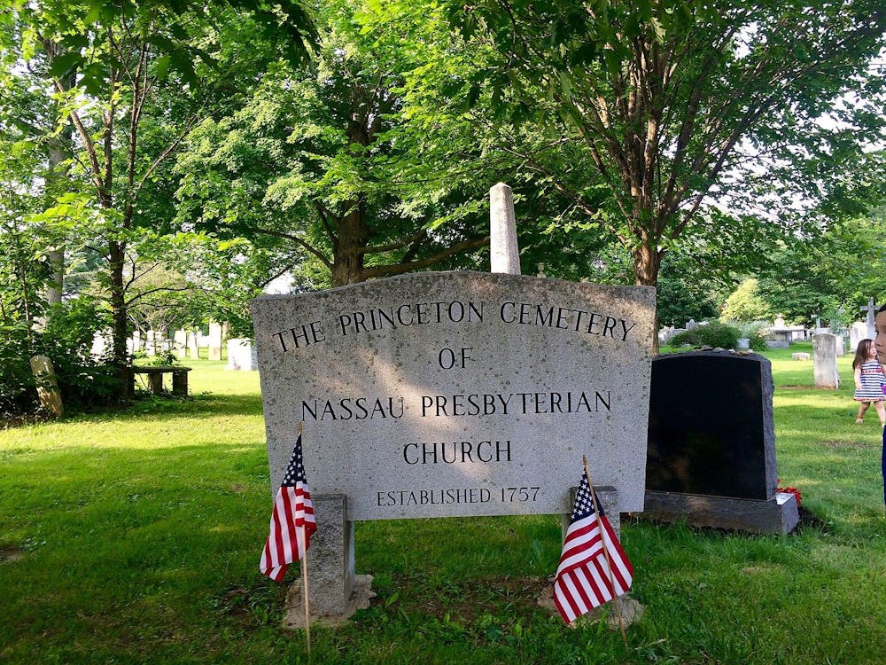 A stone sign engraved with "The Princeton Cemetery of Nassau Presbyterian Church" in front of a background of green grass and leafy green trees. Two American flags stand before the sign.