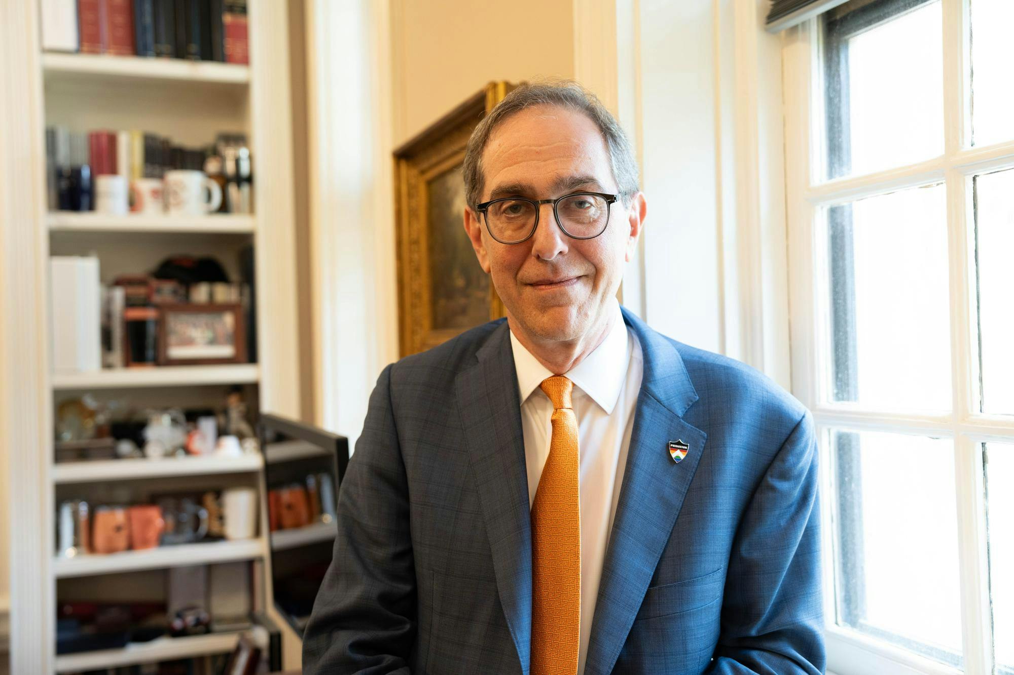 Man with blue suit and orange tie posed in front of bookshelves and window.