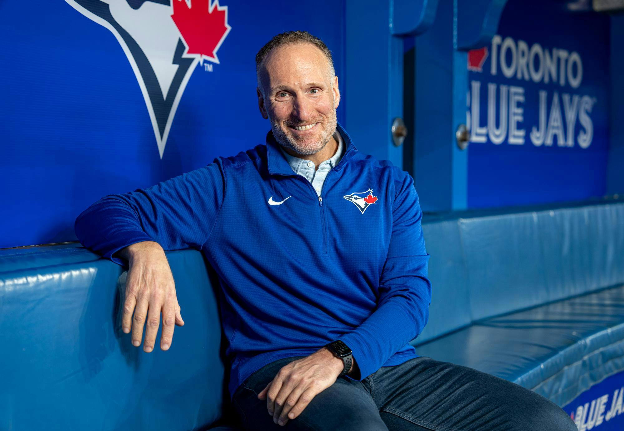 A man wearing a blue quarter zip featuring the logo of the Toronto Blue Jays and the Nike logo, posing on a cushioned seat. 