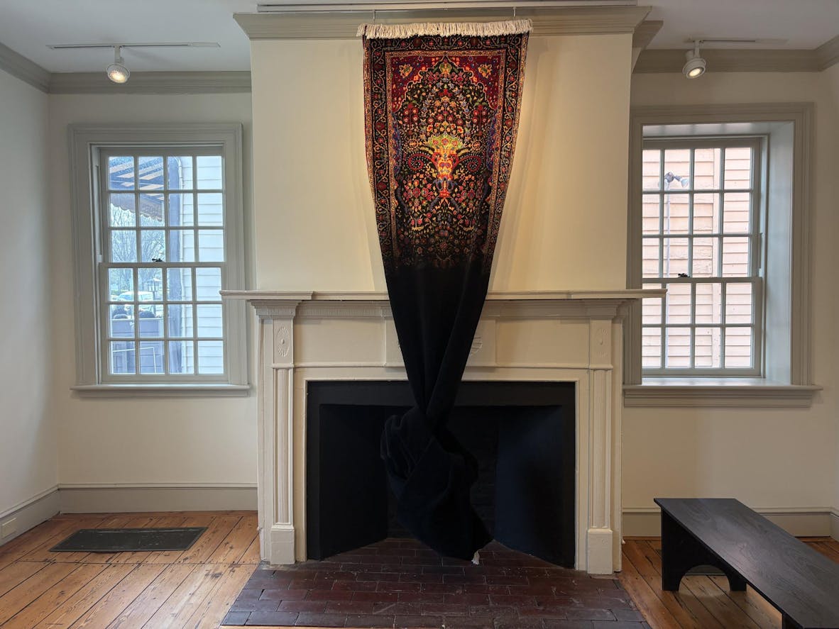 A long carpet with red, yellow, green, and blue patterns hangs against a white wall; its lower half fades into pure black and becomes a knot, then blends into the fireplace. 