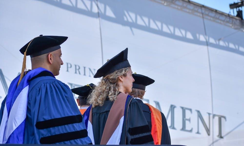 Four individuals stand with their backs to the camera wearing graduation robes in front of a large white banner