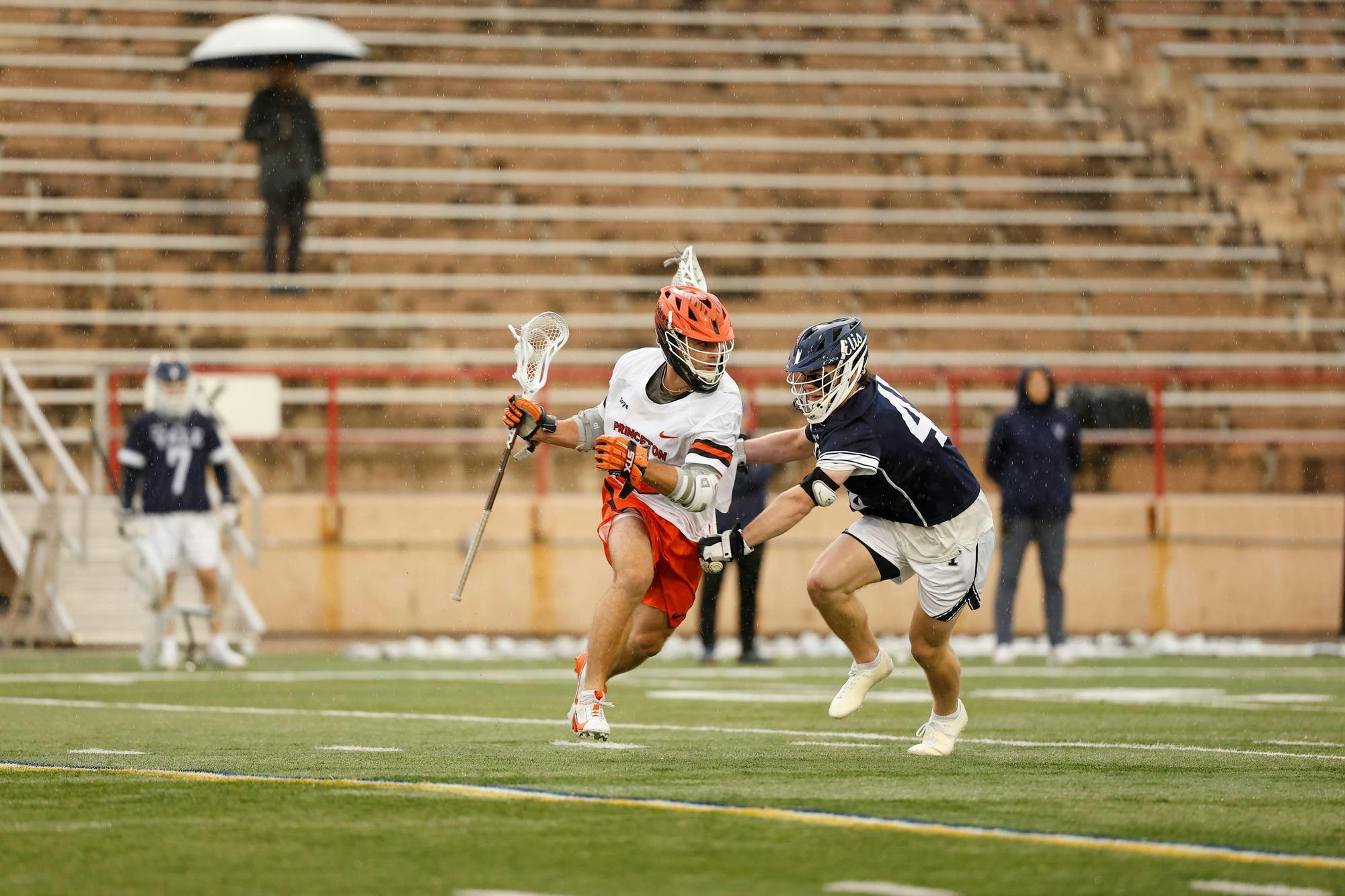 A Princeton lacrosse player in an orange helmet and white uniform drives past a Yale defender on a turf field.
