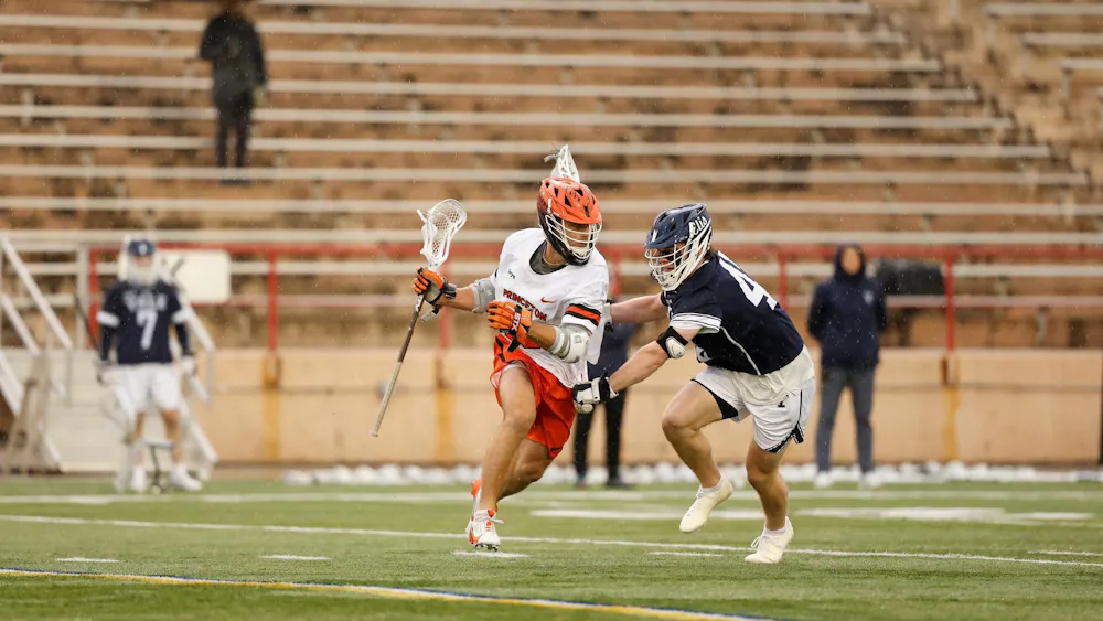 A Princeton lacrosse player in an orange helmet and white uniform drives past a Yale defender on a turf field.