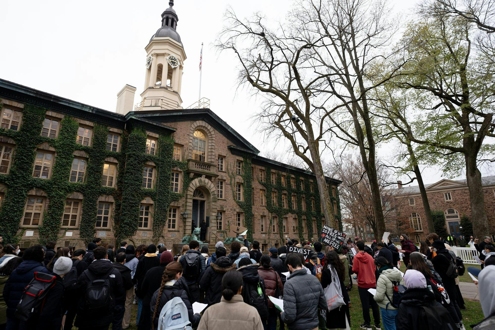 People stand in front of Nassau Hall in protest. Some hold up signs saying "Divest" and "None of us is free until all of us are free." Surrounding Nassau Hall is a white crowd-control barricade with photographers behind it. 