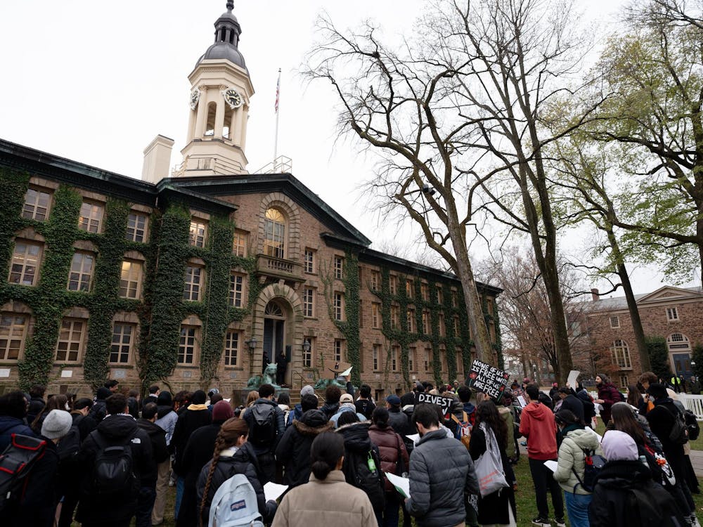 People stand in front of Nassau Hall in protest. Some hold up signs saying "Divest" and "None of us is free until all of us are free." Surrounding Nassau Hall is a white crowd-control barricade with photographers behind it. 