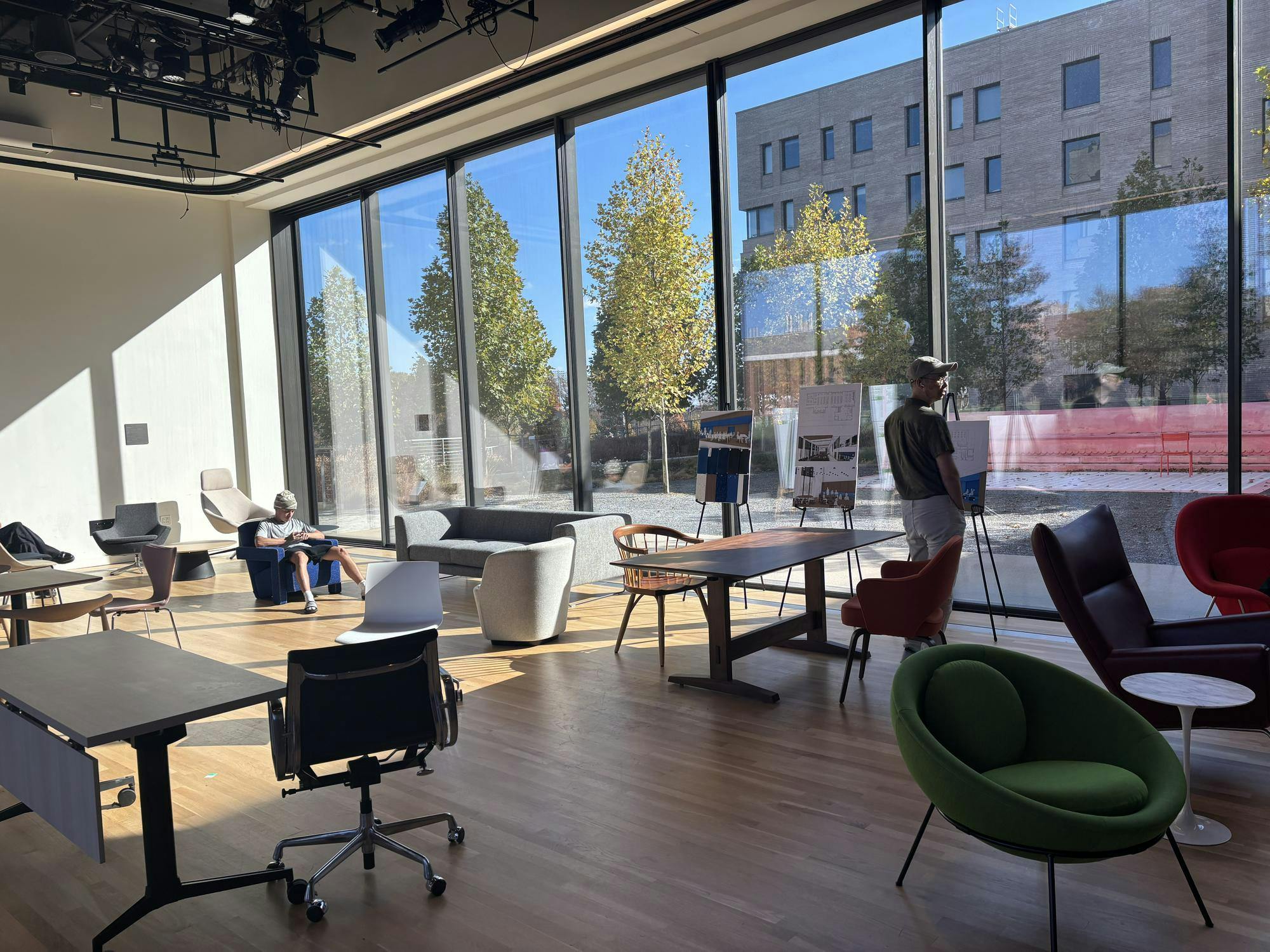A room with a glass wall with furniture in foreground. Two people are in the picture, one standing and one sitting.