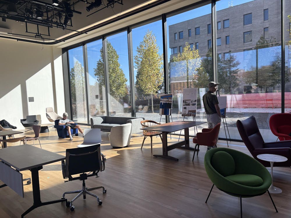 A room with a glass wall with furniture in foreground. Two people are in the picture, one standing and one sitting.