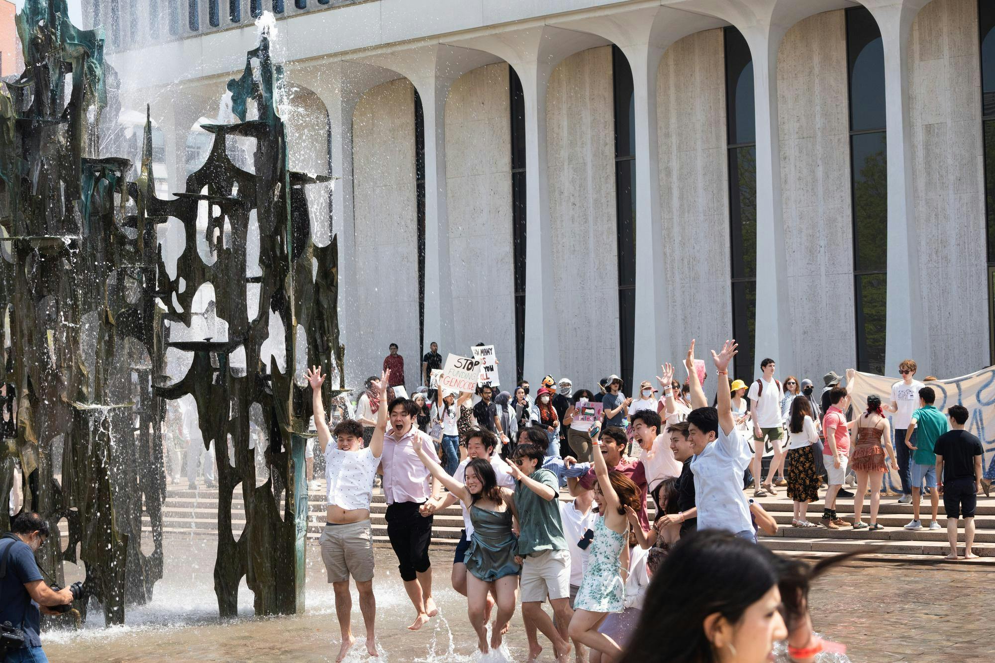 In the foreground, Lawnparties attendees wearing pastel colors jump for a photo in a fountain. In the background, protesters holding signs march by the fountain outside Robertson Hall.