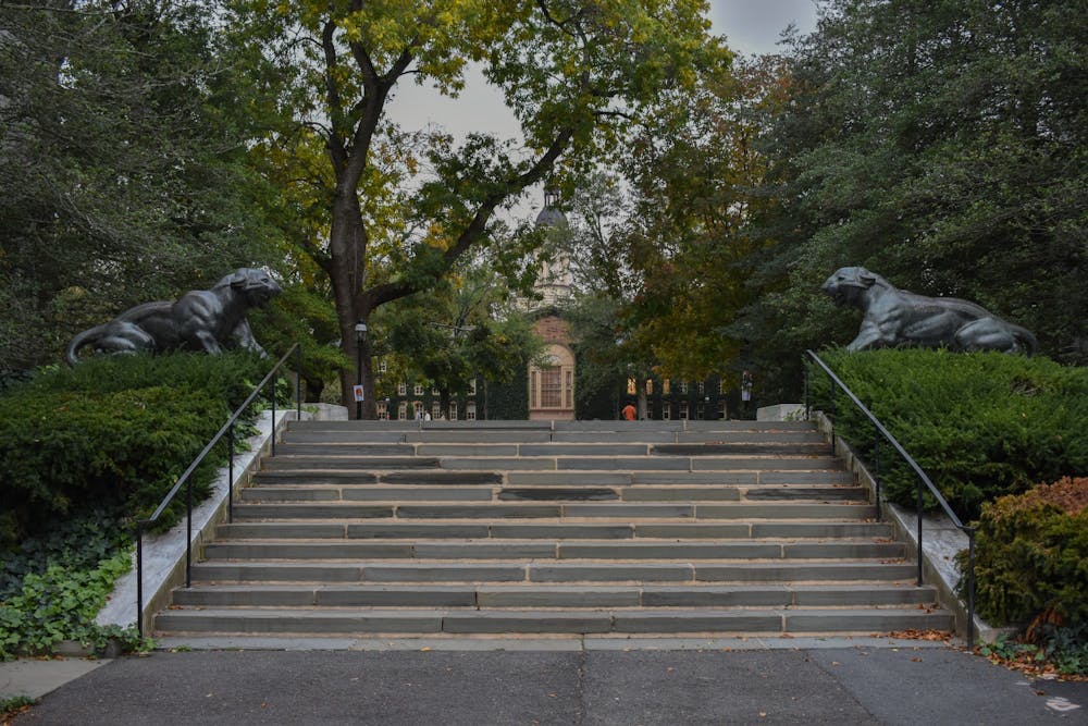 Slate steps with cement between them flanked by two shiny green statues of tigers with their mouths open facing each other.  Leafy background on an overcast day with a red brick building covered with Ivy in the background.