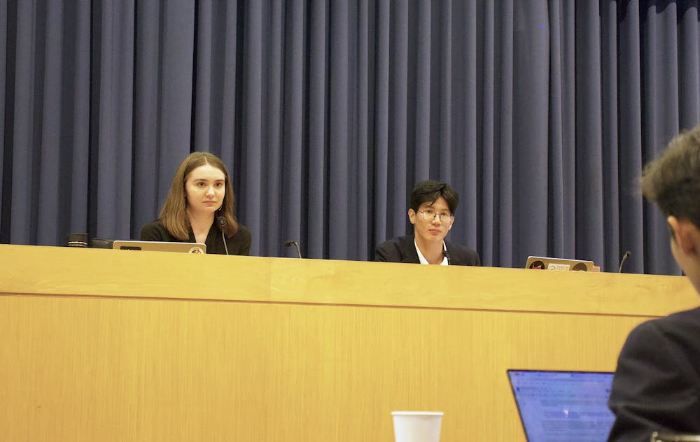 Two people sit at an elevated podium with a large navy blue curtain in the background.