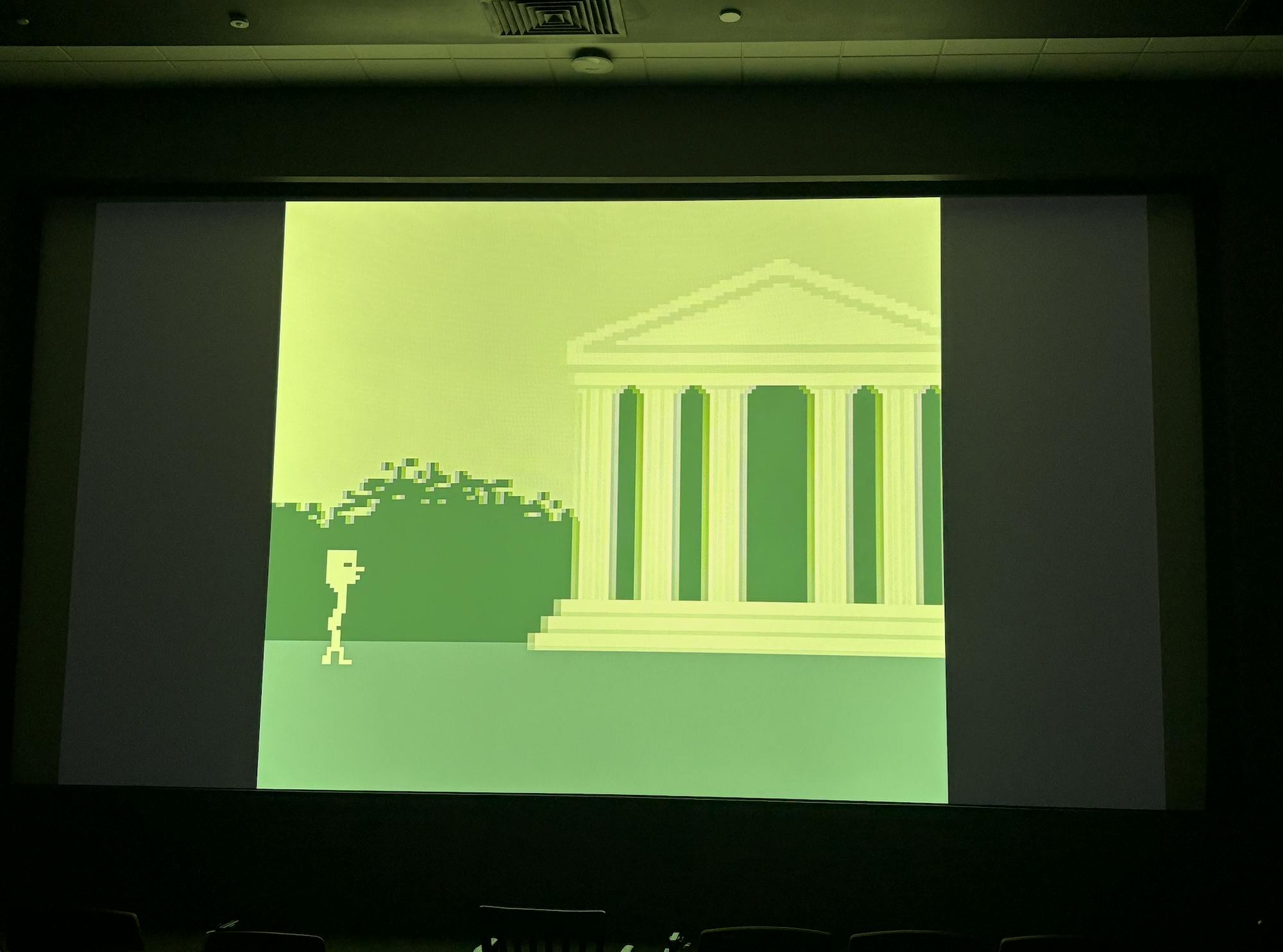 A light green, greek-style building with columns is surrounded by dark green bushes. A small, light green figure is walking towards the building. The whole scene is green and pixelated.