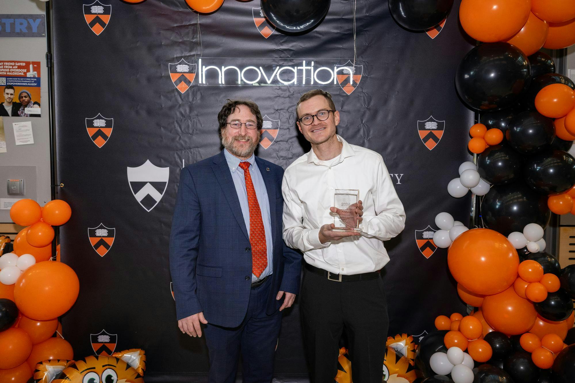 Craig Arnold stands next to Justin Silpe, who is holding a plaque for his award, in front of a banner covered in Princeton's logo. 

