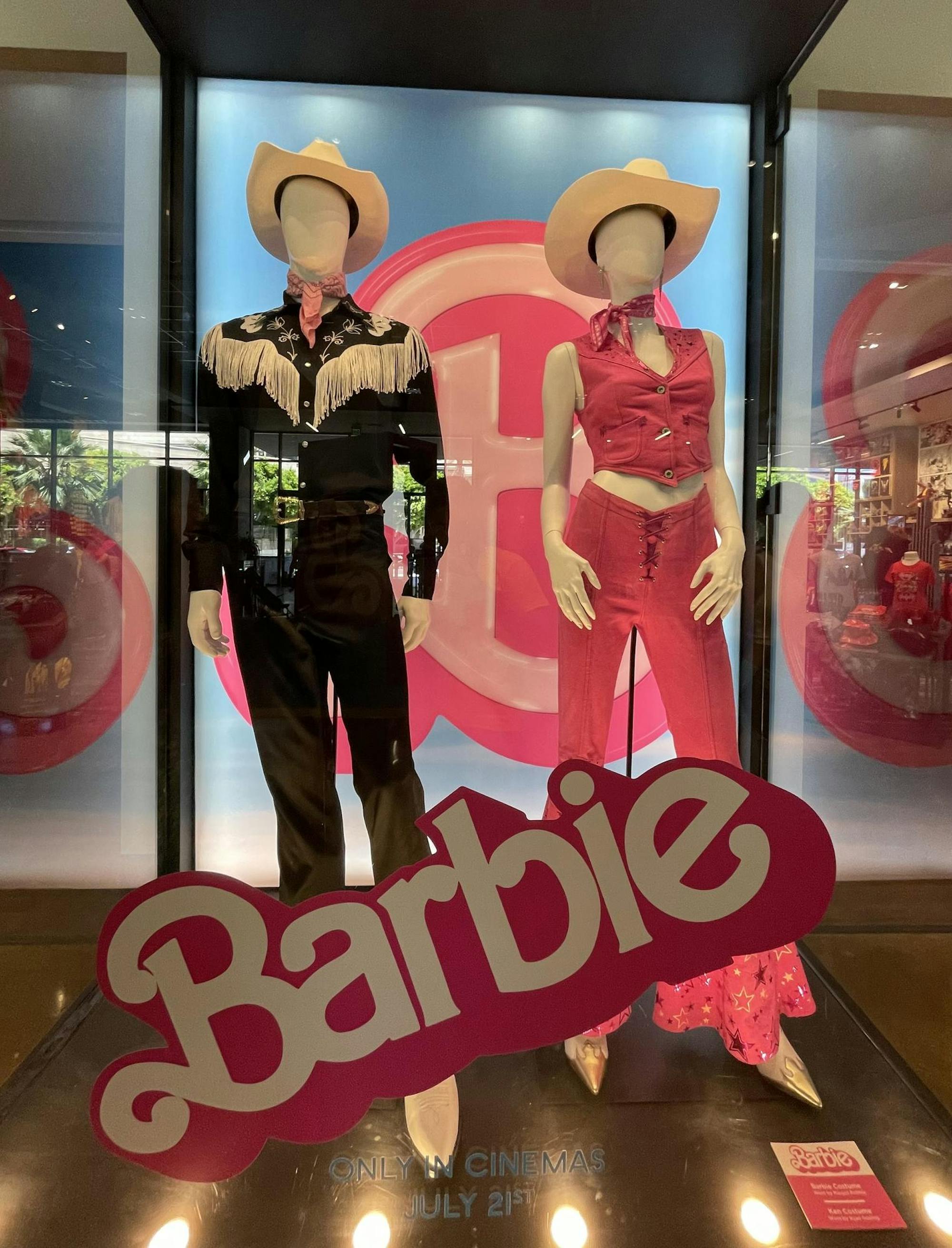 Two mannequins, one in a black outfit and one in a pink pant and shirt combo, are posed behind a Barbie display case.