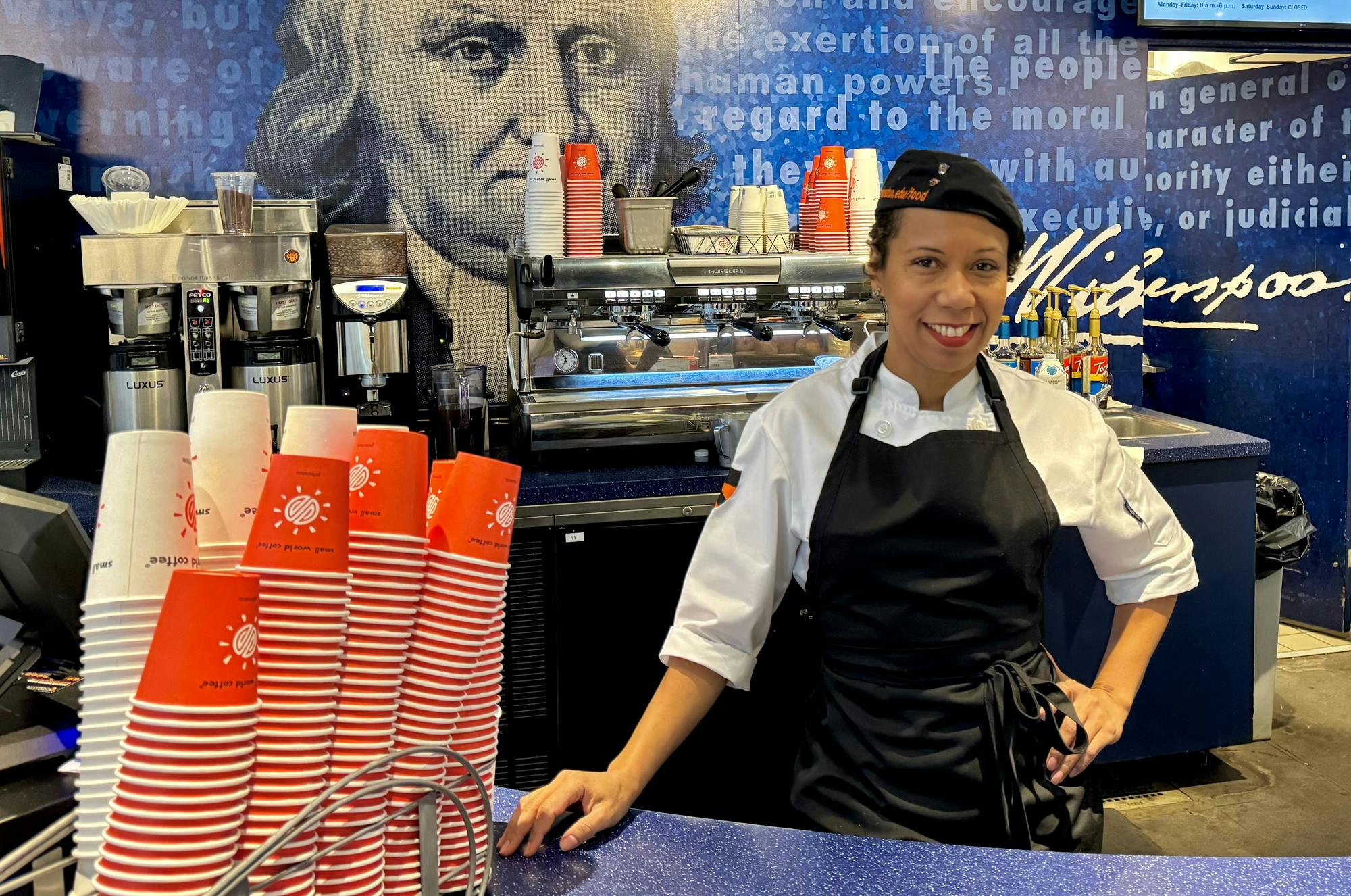 A woman in a white shirt and black apron poses in front of an espresso machine.