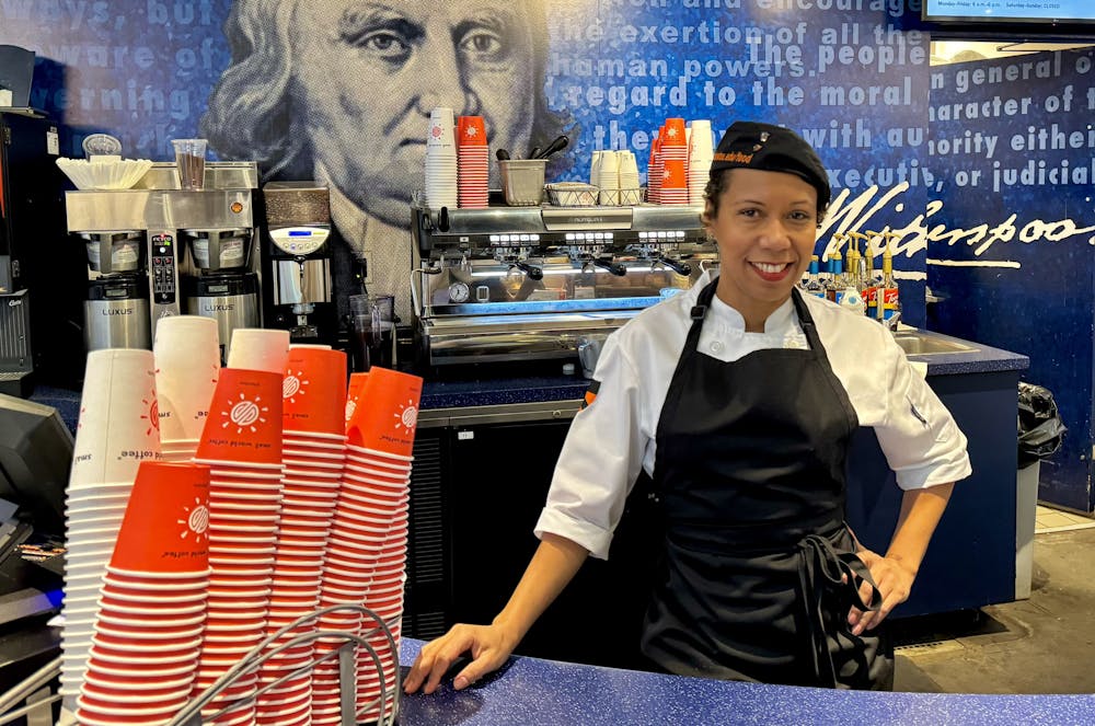 A woman in a white shirt and black apron poses in front of an espresso machine.