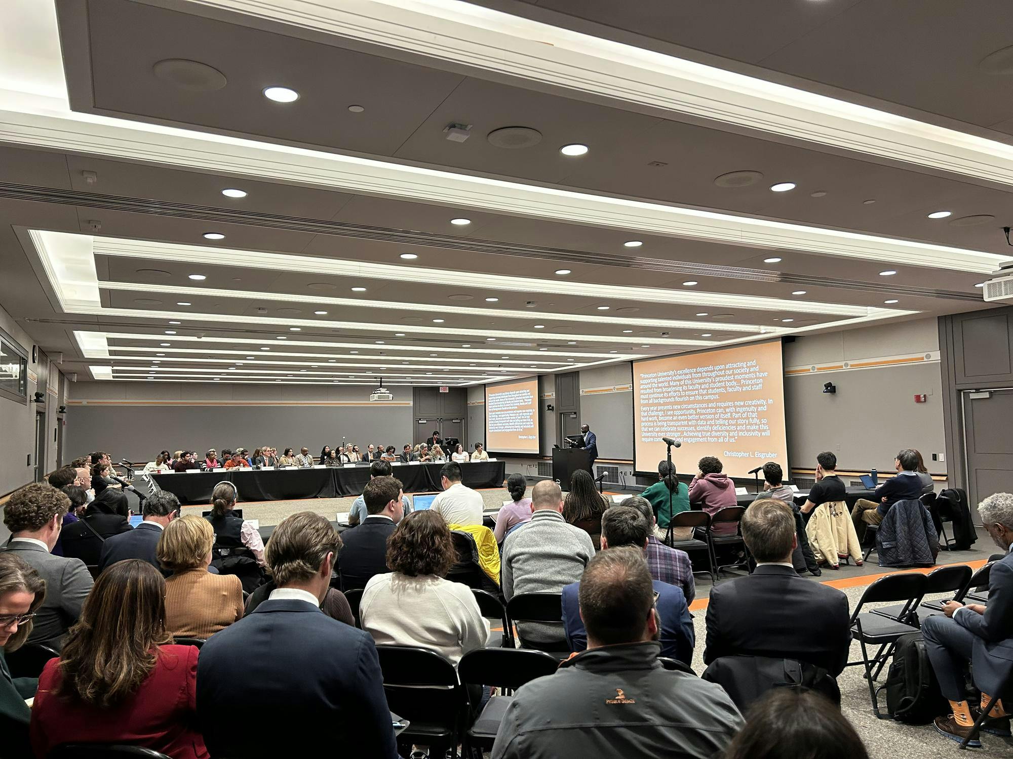 A large group of people are gathered to watch a presentation that is projected on multiple screens. They are seated at tables.