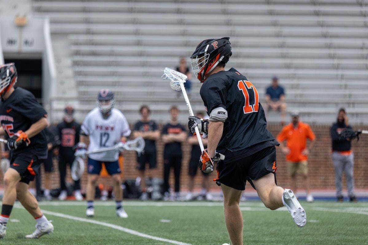 A man in a black lacrosse unfirom holding a stick with a ball in it on a field.