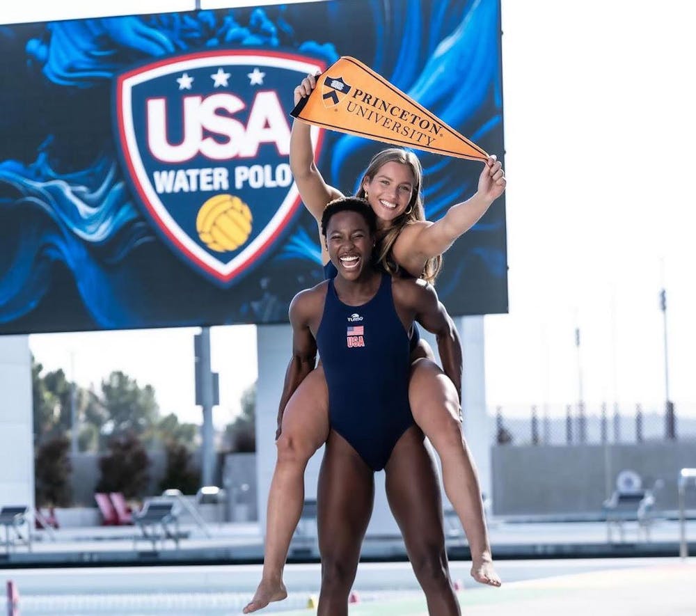 A women piggybacking on another women while holding a triangular banner saying “Princeton University” in front of a banner with the words “USA Water Polo”