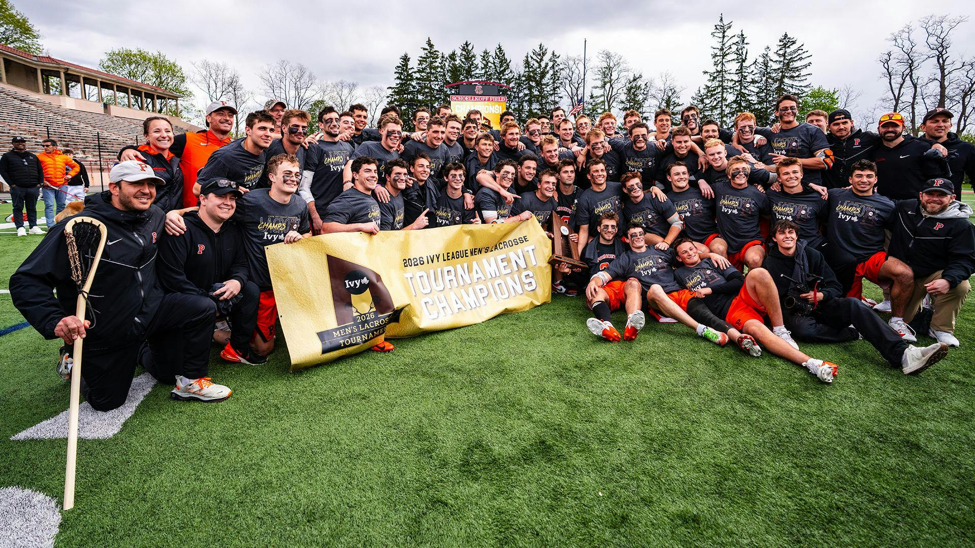 A group of players and staff pose with a tournament trophy on a grass field.