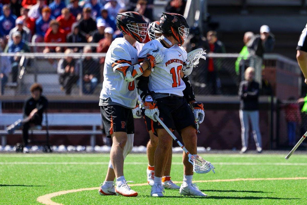 Two Princeton lacrosse players in white jerseys and black helmets stand on a sunny field.