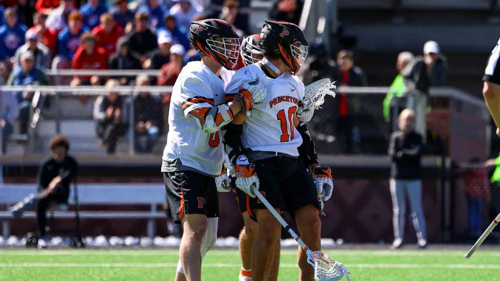Two Princeton lacrosse players in white jerseys and black helmets stand on a sunny field.