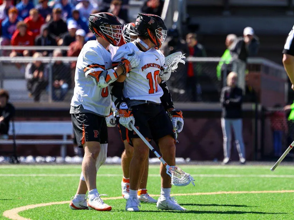 Two Princeton lacrosse players in white jerseys and black helmets stand on a sunny field.