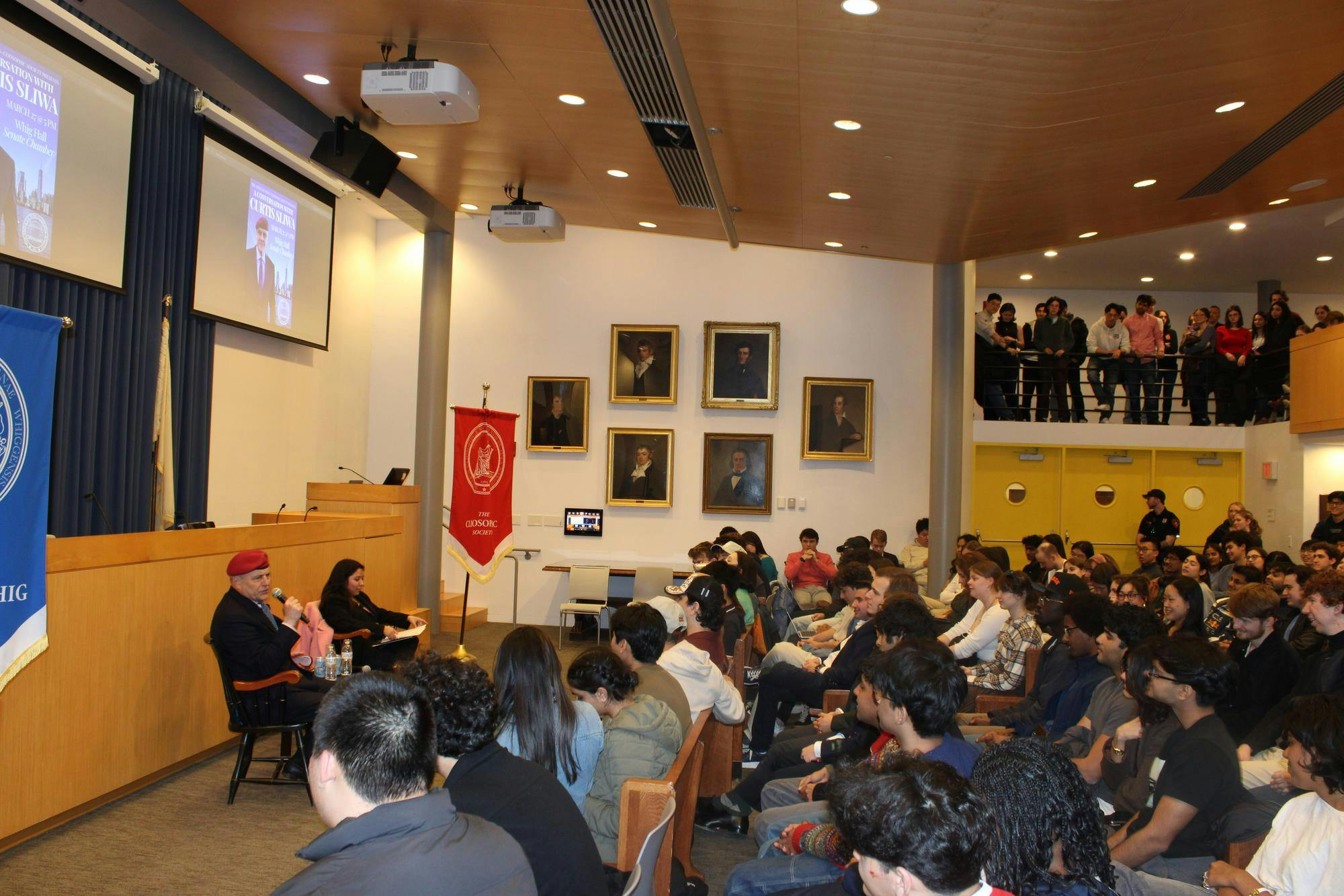 Man in a red hat speaks out at a crowd of students, holding a microphone up to his mouth. 