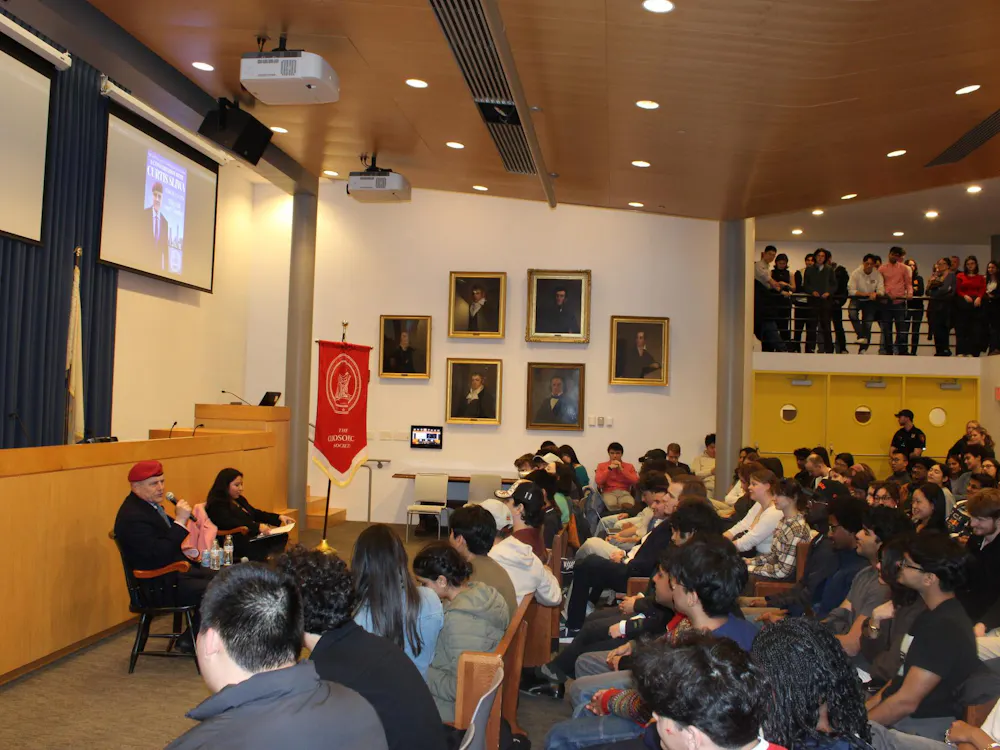 Man in a red hat speaks out at a crowd of students, holding a microphone up to his mouth.