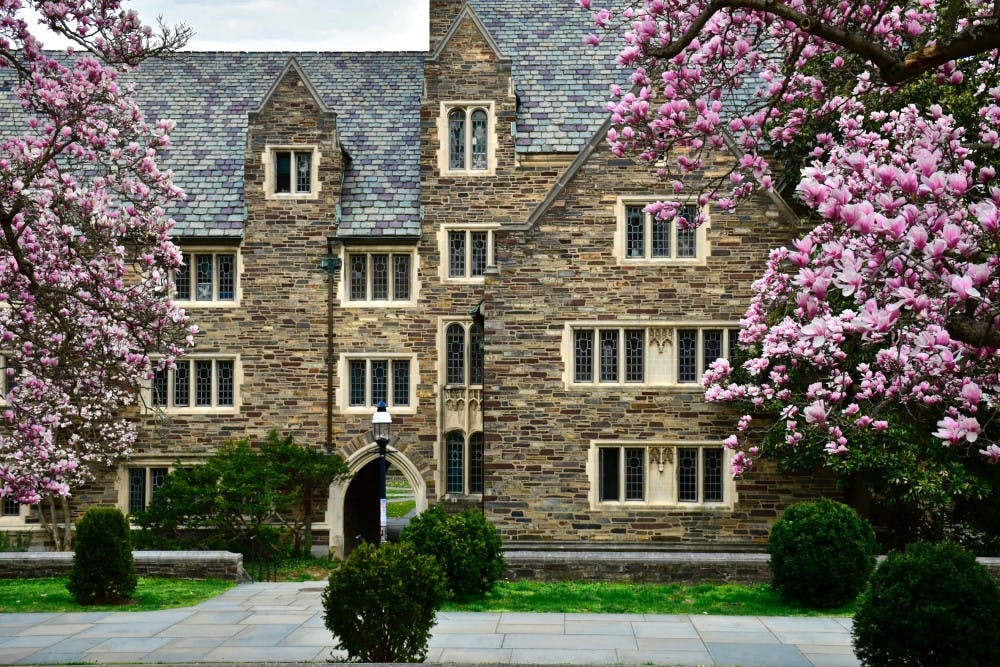 A photo of a gothic building in springtime on an overcast day. The building has a dozen windows, and trees with pink flowers surround the building.&nbsp;