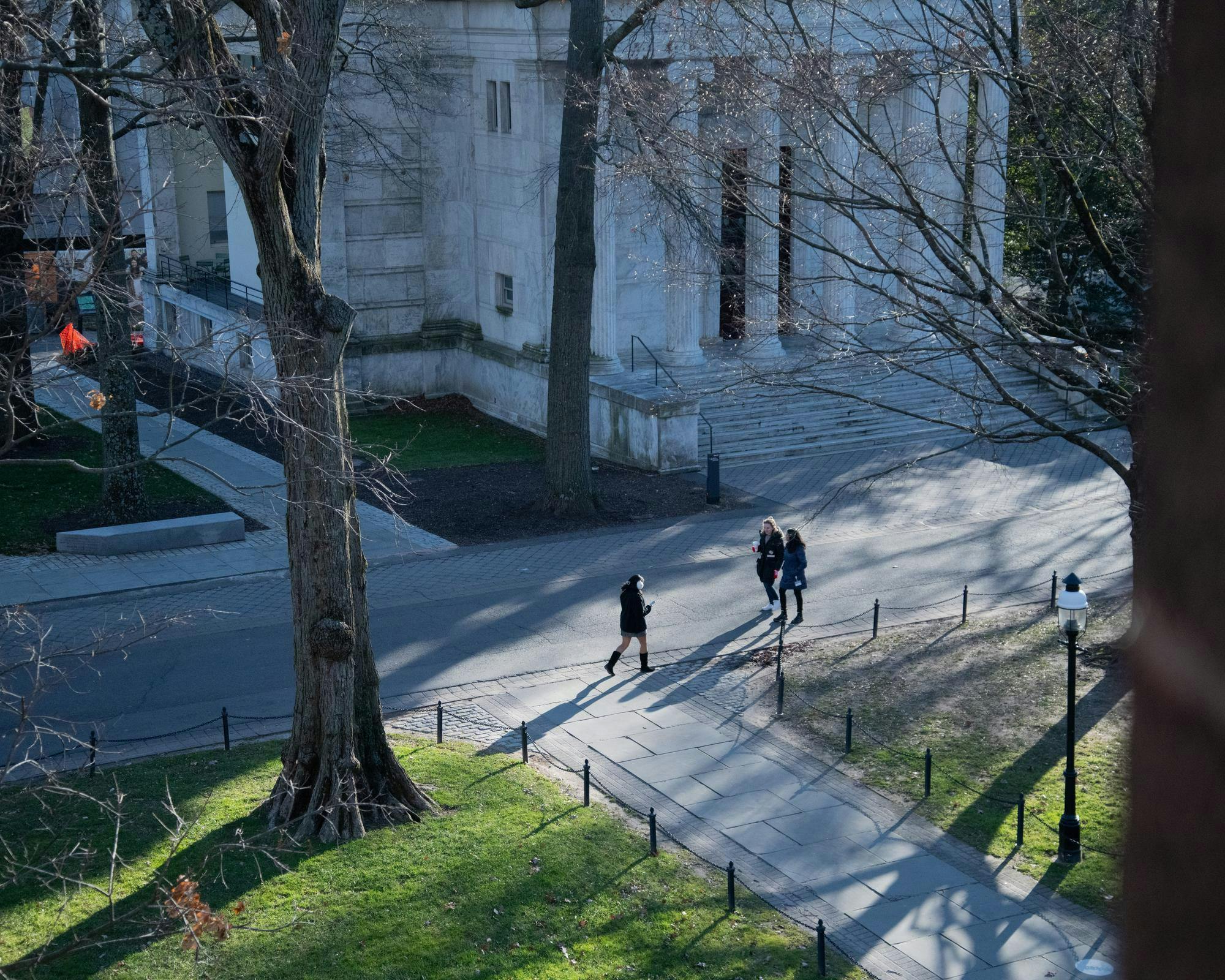 Students walking near white building with columns, surrounded by trees. Image taken from a high angle. 