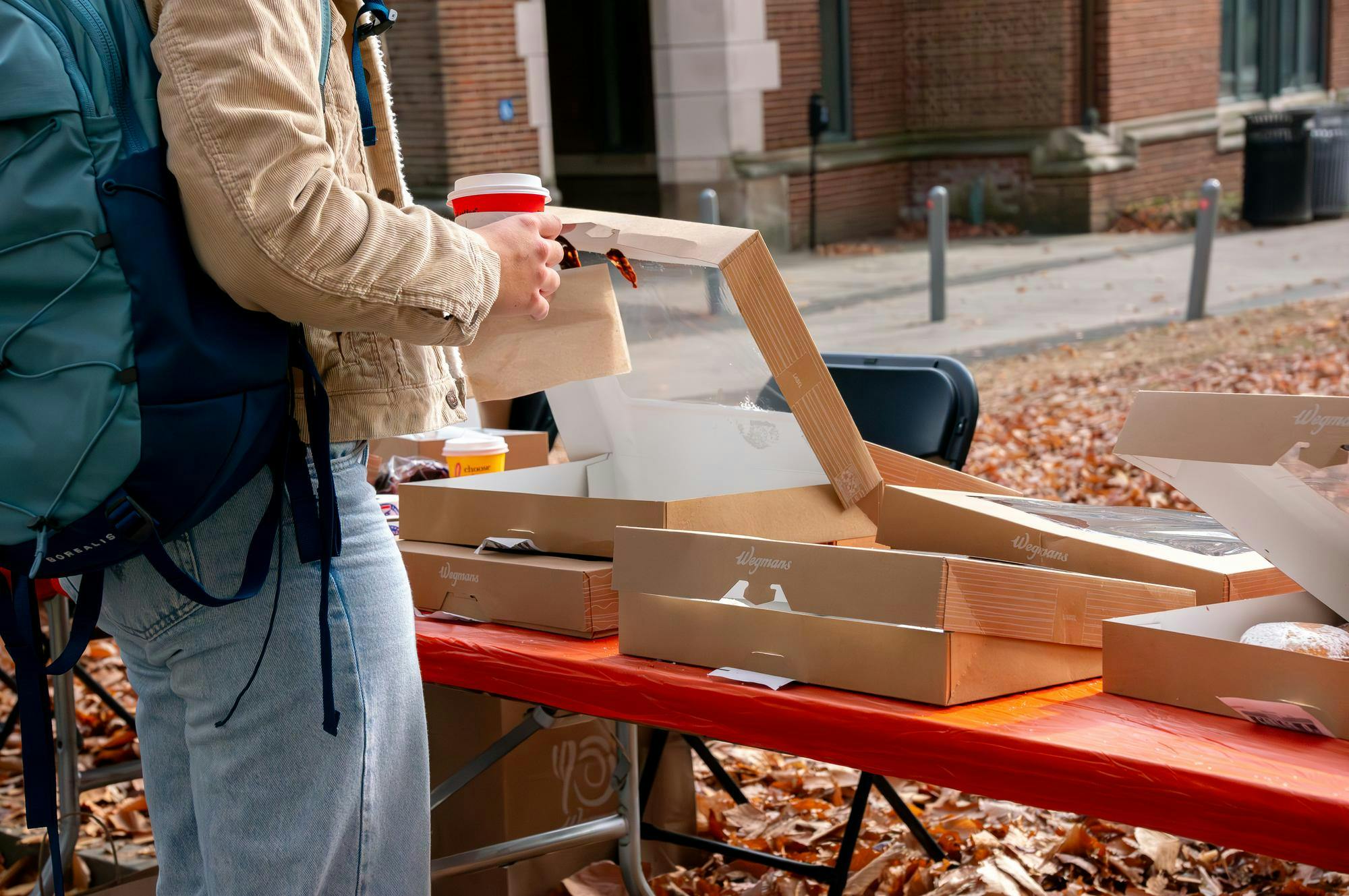 Stacks of donut boxes sit on an orange table outside. An individual is reaching into one of the boxes for a donut.