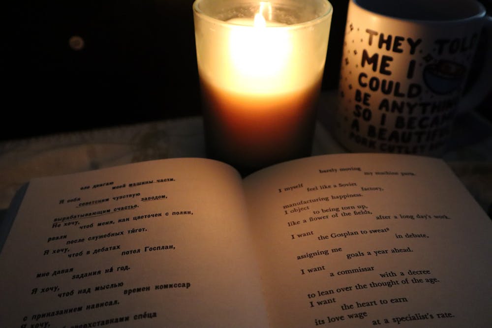 By candlelight sits an open book. On the left is Mayakovsky’s poetry in Russian and on the right, the English translation. Next to the candle is a mug with the following text written in black bubble letters "They told me I could be anything, so I became a [unintelligible text]"