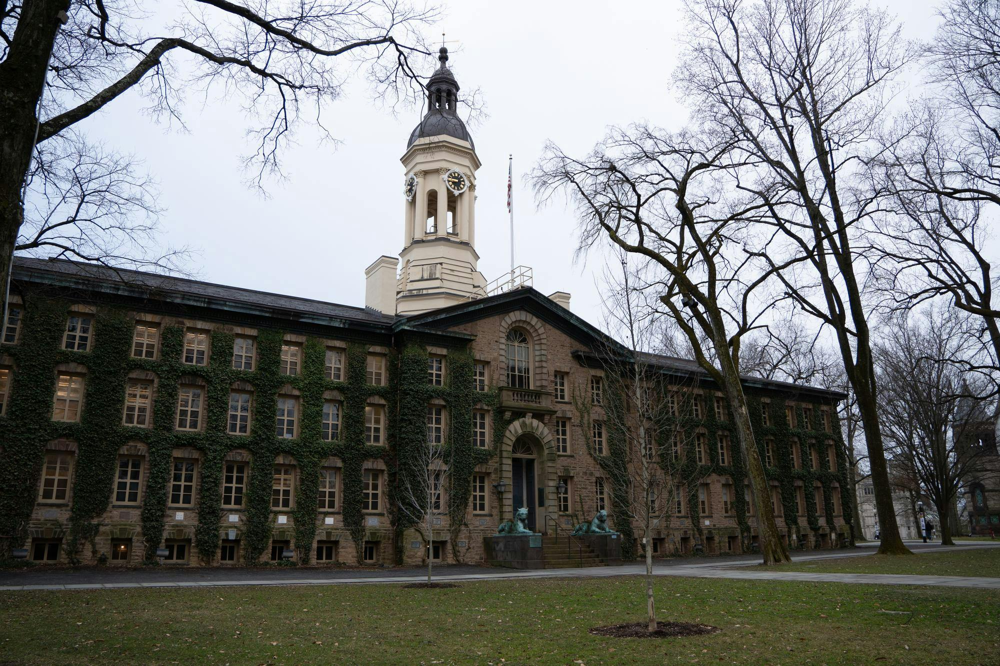 A brick building has ivy growing up the sides of it. There is a white bell tower and clock out the top.