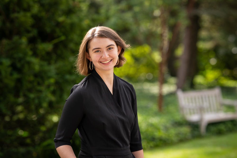 Person with short to mid length brown hair wearing a black blouse stands in front of a background filled with greenery.