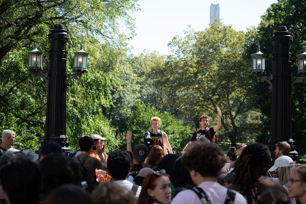 Two people, one holding a megaphone, stand in front of a crowd of people holding signs in a park.