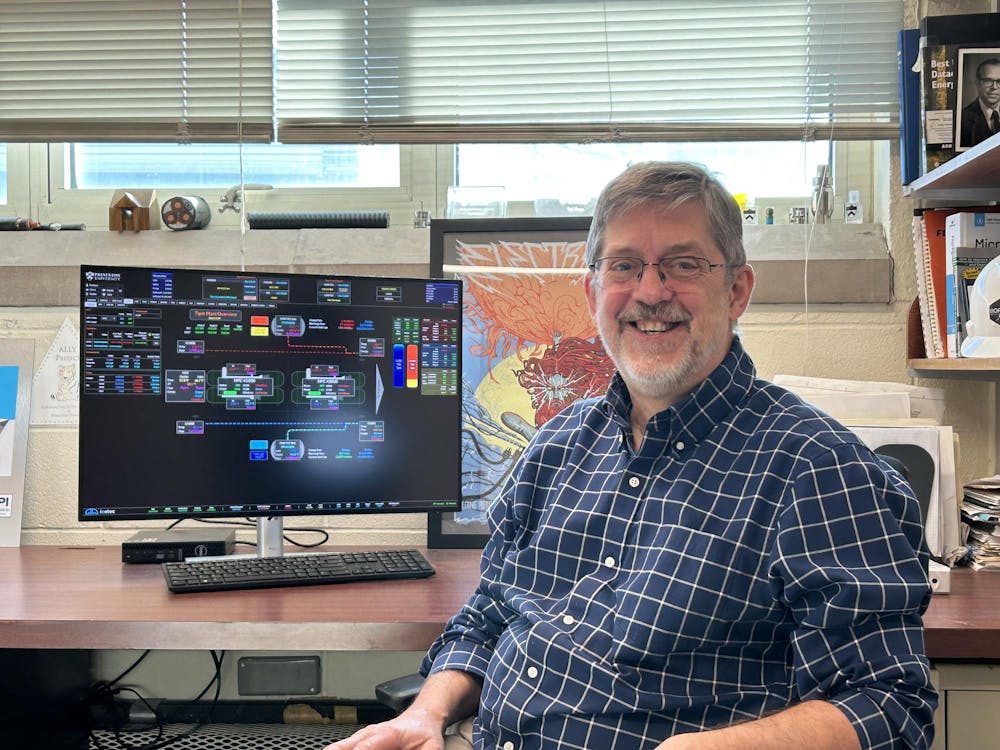 A man sits smiling in front of a desk with a computer and windows in the background.