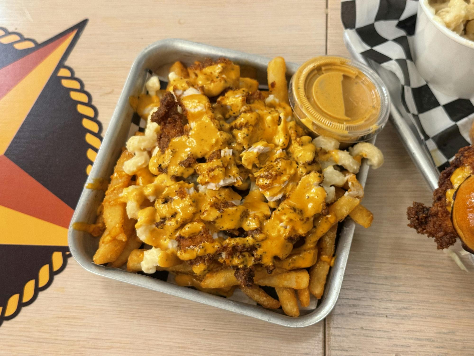 A small grey trey is lined with black and white checkered paper. There are a large pass of long, rectangular fries. The fries have a chunky mix of orange and yellow sauce and light yellow mac and cheese. There is a small transparent container with more orange colored sauce.