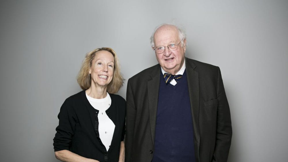 Two caucasian individuals -- one man and one woman -- pose next to each other together for a photo. The woman is on the left side wearing a sweater and the man is on the right side wearing a sweater vest, bowtie, and suit jacket.