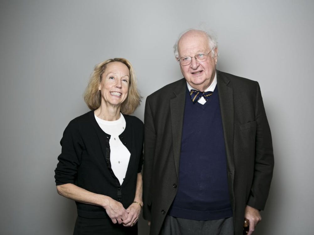 Two caucasian individuals -- one man and one woman -- pose next to each other together for a photo. The woman is on the left side wearing a sweater and the man is on the right side wearing a sweater vest, bowtie, and suit jacket.