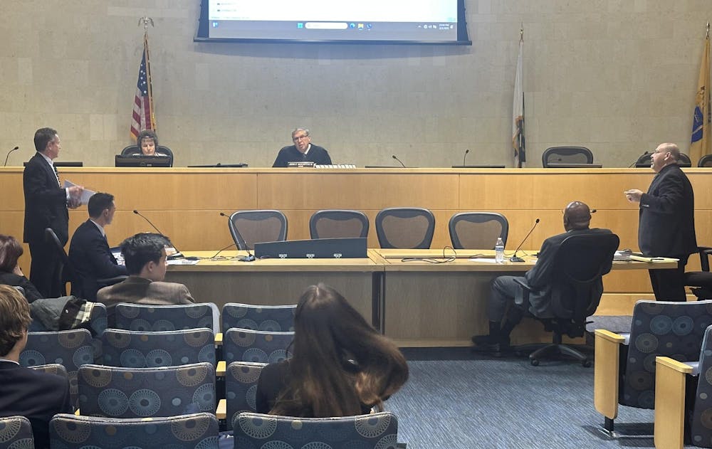 The photo shows a courtroom. A judge sits in the background on an elevated surface. At a large table in the middle, four people are gathered. Multiple people are sitting in the audience.