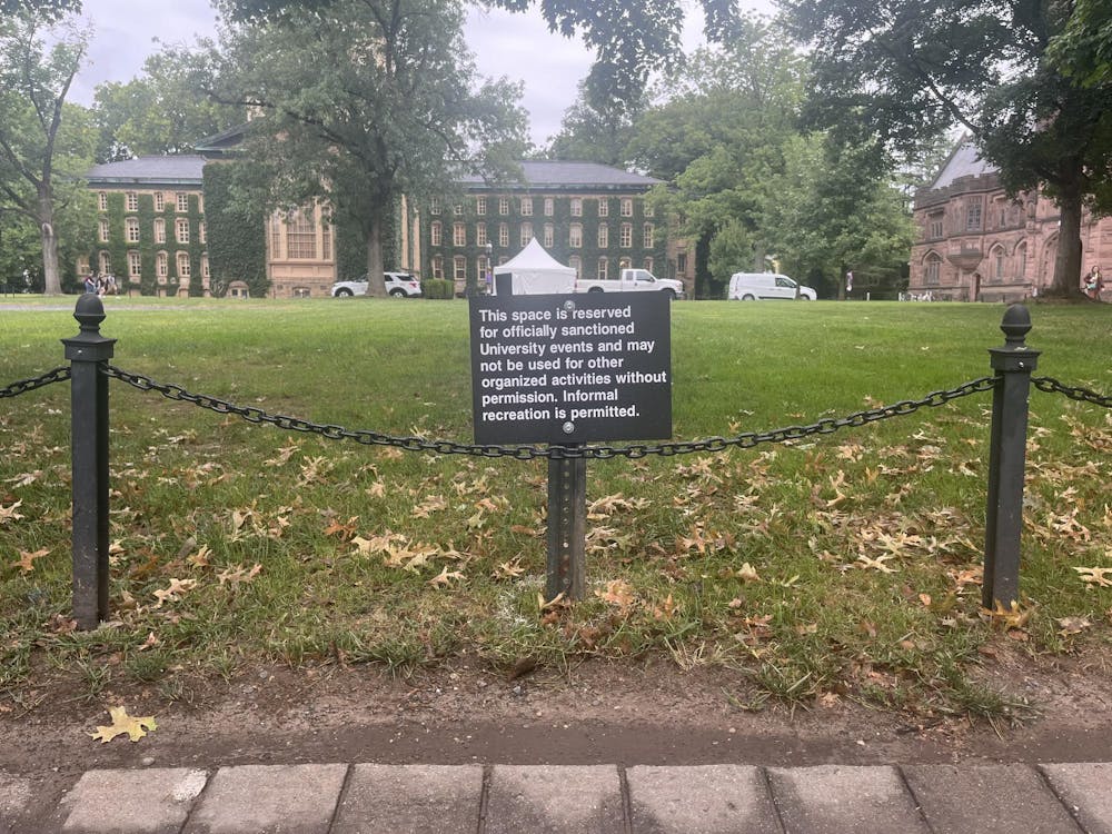 A small black sign reads, “This space is reserved for officially sanctioned University events and may not be used for other organized activities without permission. Informal recreation is permitted.” A green lawn behind the sign is partitioned by a black chain rope. A large, ivy-covered building stands in the background.