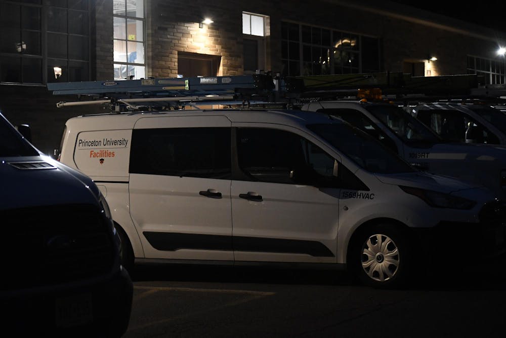 A white Ford Transit Connect with a "Princeton University Facilities" sticker on the side sits parked behind Baker Rink. It is night and there is dim lighting.