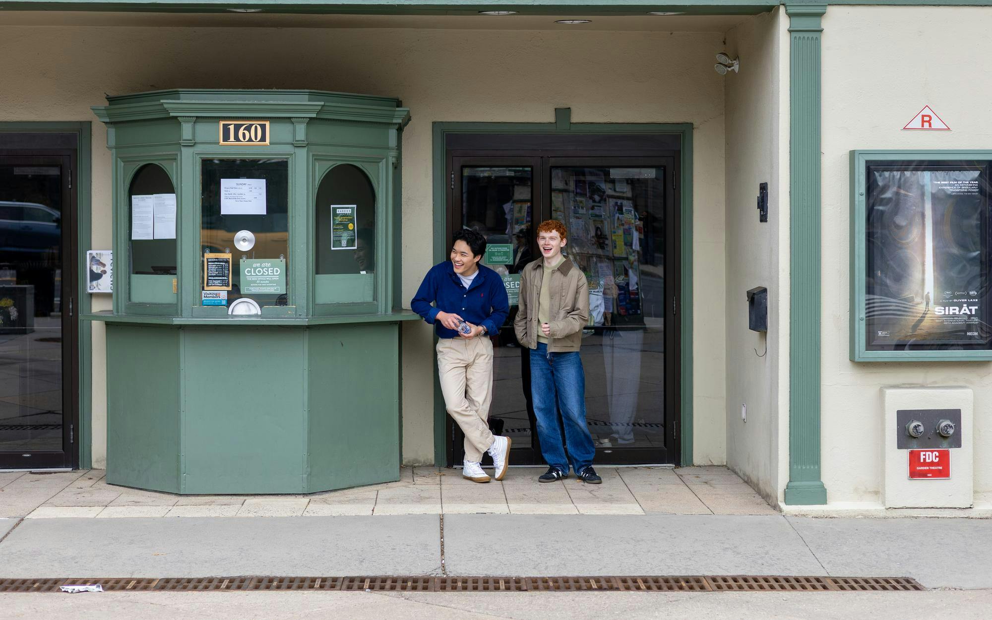 Two students in multicolored outfits stand in front of a building that has a green and white facade.