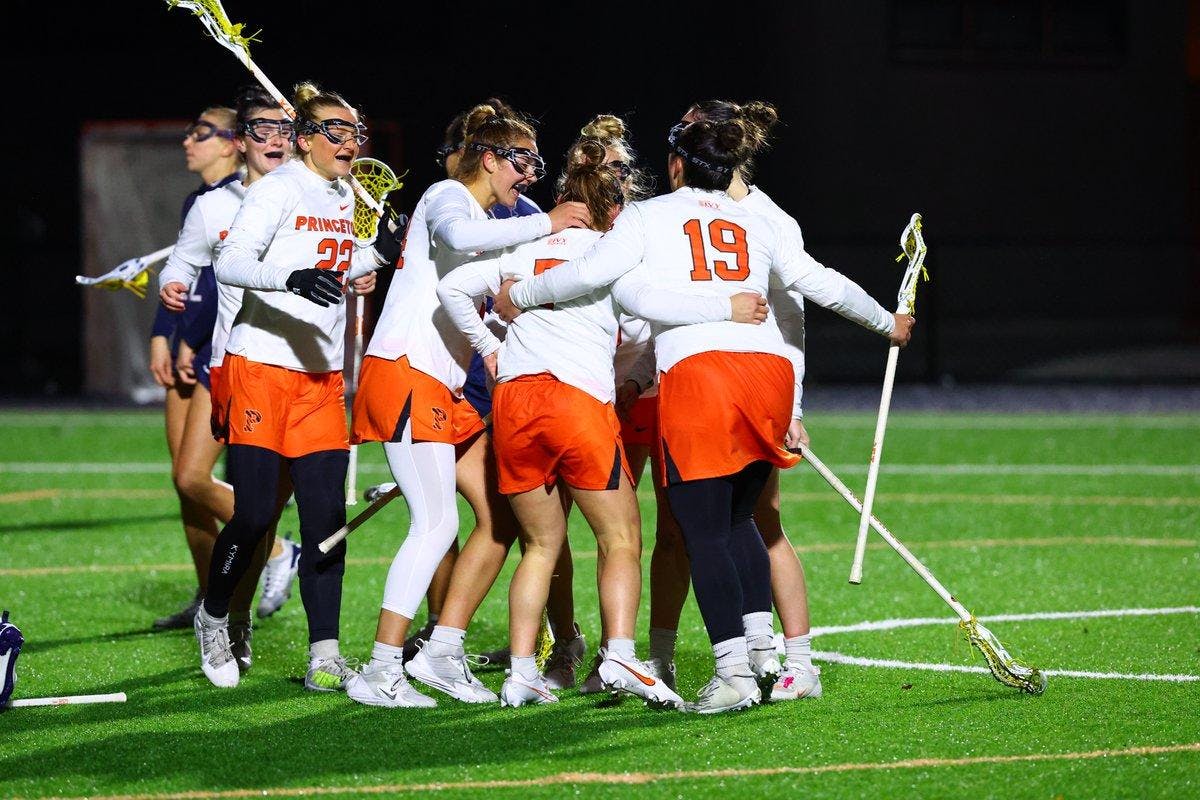 Huddle of Princeton lacrosse players with white jerseys and orange shorts gather on the green field. The girls are hold lacrosse sticks. 