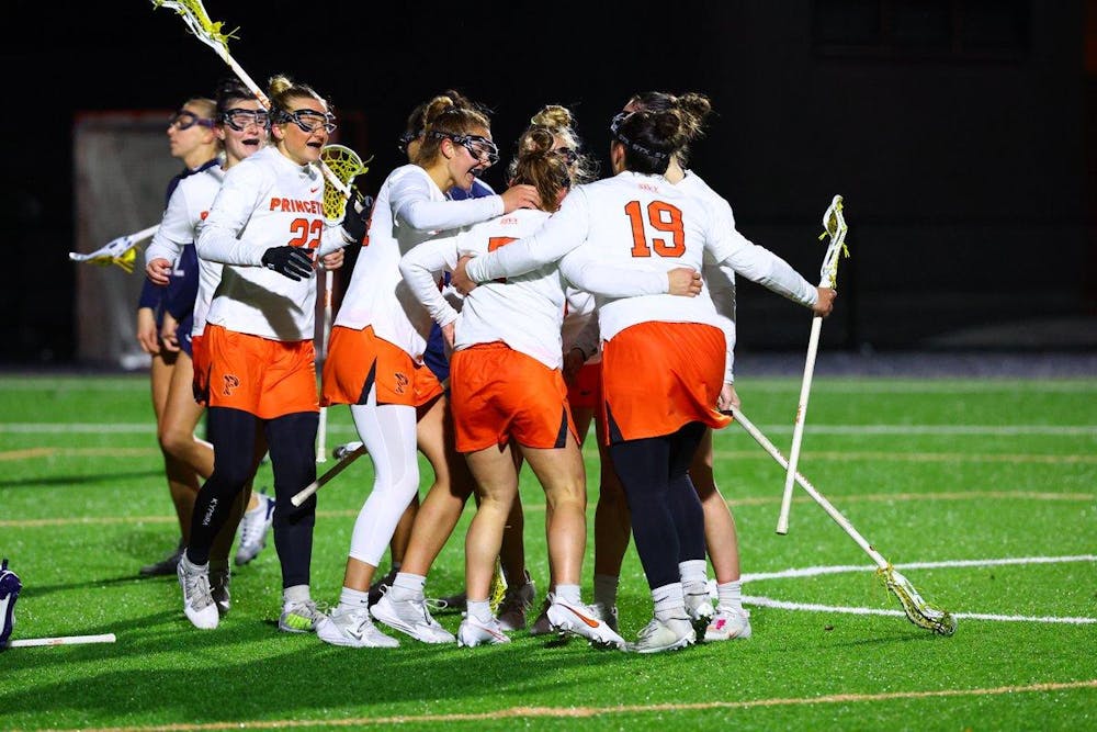 Huddle of Princeton lacrosse players with white jerseys and orange shorts gather on the green field. The girls are hold lacrosse sticks. 